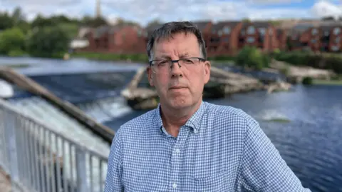 A man in a blue checked shirt stands by the River Exe at Trews Weir in Exeter. The weir is in the background and the water flowing over it is subdued. 