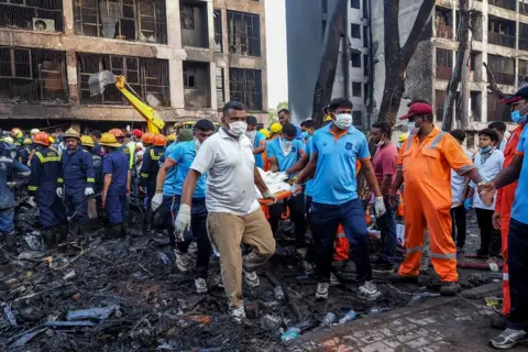 AFP via Getty Images Rescuers wearing a variety of clothing - including fire officials - retrieve a body covered by a blanket, on a stretcher, from the ruins of the medical college on Thursday