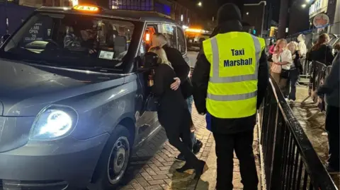 Peterborough City Council A couple speaking to a taxi driver tough his taxi window, with a person wearing a green high-visibility jacket with "taxi marshal" written at the back standing in front of the taxi.
