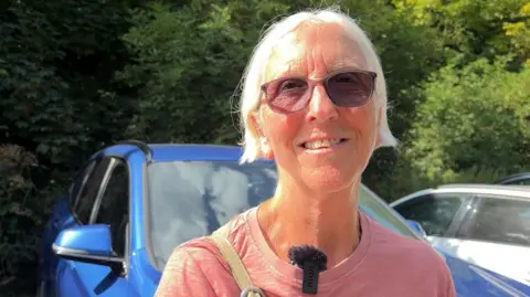 A woman with short grey hair, sunglasses, and a pink t-shirt smiles as she discusses Ros Canter's medal win. 