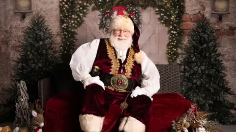 Danielle Axworthy Allan Evans, in a full Santa suit, sat on a velvet red throw with Christmas trees in background and woodland animal ornaments at his feet
