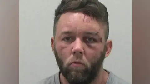 A mugshot of Ross Neville. He has brown hair and a beard. He has cuts across his left eyebrow and on his forehead.