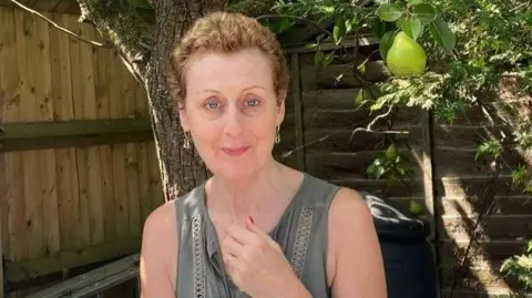 A woman stands in a sunny garden beside a wooden fence, surrounded by greenery. A large green pear hangs prominently from a tree branch overhead.
A black cylindrical container sits on the ground nearby. A metal ladder leans against the fence, suggesting recent or ongoing yard work.