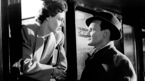 Getty Images A black and white scene from Brief Encounter showing Celia Johnson in a heavy coat leaning from the window of a train to look into the face of Trevor Johnson who is standing on the platform wearing a dark coat and hat. They are holding hands