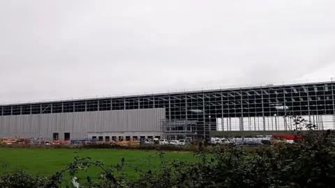 The image shows a giant warehouse being built on land next to the Severn Estuary but close to houses in Pilning. 