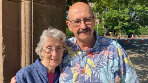 Margaret and Phil are photographed smiling in the spring sunshine in Sefton Park. Margaret has grey hair and glasses, while Phil is bald, has a moustache and also wears glasses.