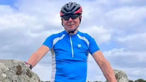 John Lyttle wearing a blue and white short-sleeved athletic top, sunglasses and a grey bicycle helmet. He is standing outdoors on a sunny day, with lots of fluffy clouds behind him, leaning against a rock covered in lichen and smiling at the camera.