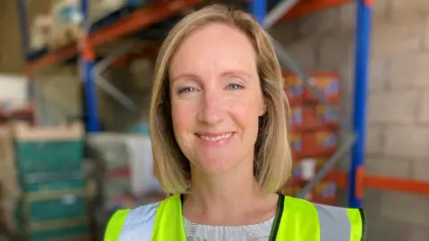 A woman with blonde hair wearing a high-vis yellow jacket standing in a food warehouse