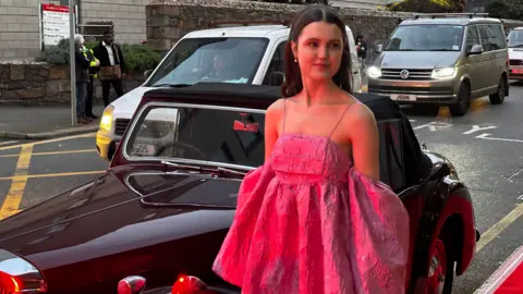 Chloe Sweetlove wearing a voluminous pink dress stands in front of a the Bergerac red triumph roadster outside Jersey Opera House. Several vehicles, including a white van and a silver van, are visible in traffic behind.

