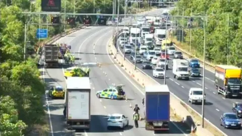 The M6 motorway in Birmingham northbound with police cars and fire engine workers surrounding lorry that has caught fire. The southbound shows queues of cars.