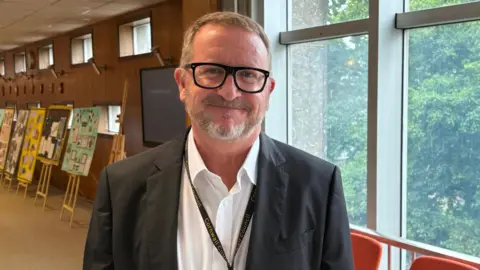 A man wearing a white shirt and blazer smiles at the camera. He is standing indoors in front fo a large glass windows with trees visible. 