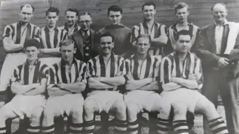 A black and white photo of a football team, its club chairman, and trainer, posing for a team photo. There are five players sitting in the front row, while the other six players, as well as the chairman just left of centre and the trainer on the right, stand behind them. The outfield players wear white-and-dark striped jerseys, with white shorts and hooped socks seemingly of the same colours as the jerseys. The goalkeeper wears a dark top, while the chairman, who has glasses, wears a suit, tie and a patterned waistcoat. The trainer on the right wears a dark jacket and trousers, a white shirt with no tie, and has a towel over his left shoulder.