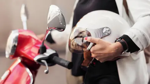 A woman in a cream jacket and black outfit holds a white helmet in one hand, and a moped handlebars with her other hand. The moped is red and has chrome lights.