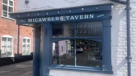 Neve Gordon-Farleigh/BBC A blue shop front at the corner of a white-brick building. The shop front has "MICAWBERS TAVERN" written above its window. 