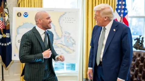 The White House Conor McGregor in a green suit in the Oval Office with blonde-haired Donald Trump, who is wearing a navy suit.