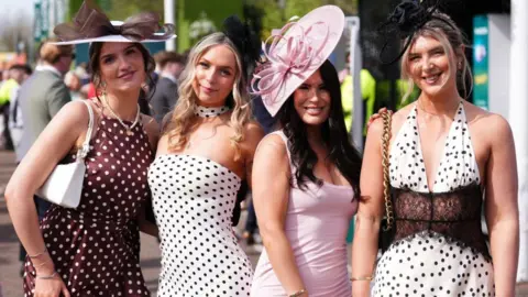 PA Media Four women stood in a line smiling at the camera. Three are weraing polka dot dresses and a fourth is wearing a lilac dress.