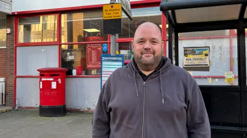 A man with a beard and a grey hoody stood outside a post office with a post box behind him