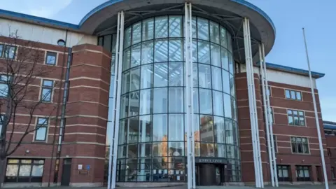Front view of a modern red-brick justice centre building with a large curved glass atrium entrance, supported by tall white columns and topped with a sweeping roof canopy. The building spans several storeys, with rows of windows on either side of the central glass façade under a clear daylight sky.
