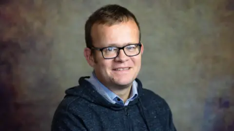 University of Reading Professor James Reade looking at the camera has dark hair and eyes, is wearing black framed-glasses. He has a blue shirt on and a navy zipper jumper over the top. 