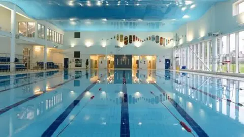 A large swimming pool with light blue tiles on the bottom and dark blue tiles separating lanes. There are glass windows on the right  and a viewing area with dark blue chairs on the left