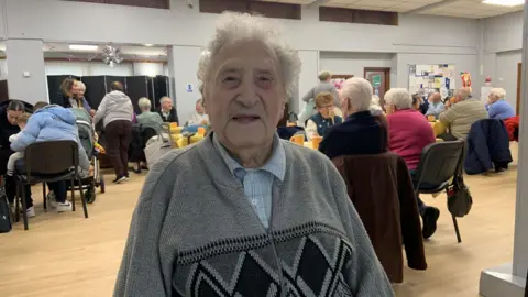 An elderly man with grey hair is wearing a grey zip up cardigan and blue checked shirt. He is sitting in a community centre hall with people seated in the background