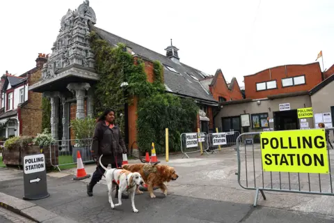 Peter Nicholls / AFP via Getty Images A white and brown dog are walked by a person wearing a coat with long hair past two signs saying 'polling station' outside the temple against a cloudy grey sky.