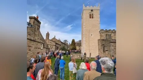 LDRS St Oswald's Church in Askrigg which is a square tower with has been lime washed, and there are a crowd of people standing around it. 