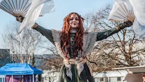 Kildare County Council A woman dressed in a Celtic-inspired costume to represent a figure of St Brigid for Kildare's Spirit of Brigid festival.  She has long, ginger hair and is wearing a green tunic and a gold belt and is holding a gold fan in each hand.