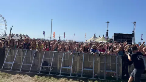 Thousands of people stand behind a barrier at Boardmasters in Cornwall. A camera man is on the right of the image. A big top and ferris wheel are in the background.
