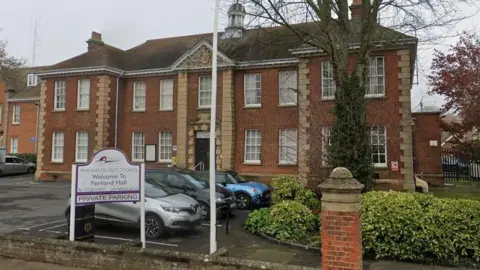 A red brick two-storey Georgian-style municipal building. It has light-coloured corner capstones, a pitched roof and a tiny cupola in the middle of its roof. There is a car park with some cars in front and a low wall with a white sign saying "Welcome to Fenland Hall"