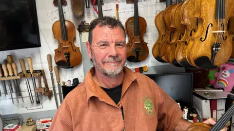 Andy Parish has short black hair and grey moustache and beard he is wearing an orange heavy shirt with open collar and a black shirt underneath, he is surrounded by violins and is holding one in his hands.