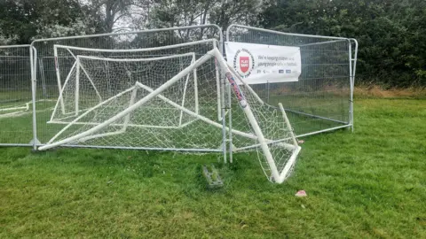 Lee Bailey A plastic goal half under tall fencing, bent over in a field with trees at the edge