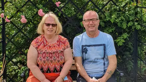 Audrey, with blonde bobbed-hair and sunglasses, wears an orange patterned top and matching orange trousers. She is sitting on a garden bench surrounded by flowers with her late husband Eddie on holiday in 2020.