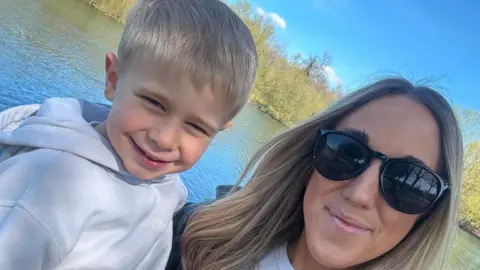 Supplied Reggie and Jade stand looking at the camera and smiling. Behind them is a lake. Jade has long blonde hair and dark sunglasses on. Reggie is wearing a white hooded sweatshirt and has blonde hair. 
