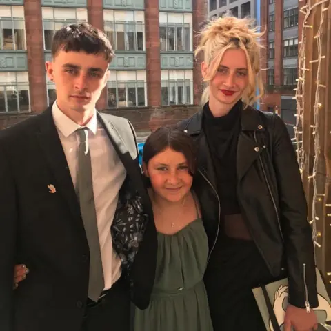 Jobson family A young man with brown hair in a black suit jacket, a white shirt and grey tie stands next to a small girl with brown hair and a green top, and a young woman with tied-up blonde hair, a black blazer and black jumper.