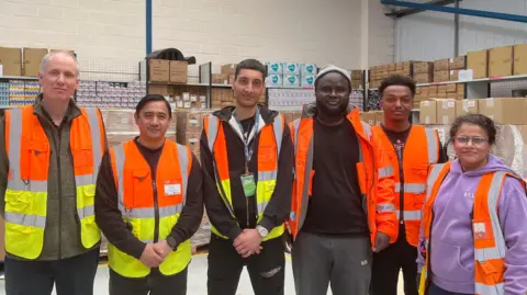 A group of people with high-vis orange and yellow jackets looking into the camera. There are six people looking into the camera they are all slightly smiling. Behind them is a warehouse full of boxes. 