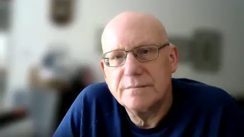 A bald man in dark glasses wearing a dark blue t-shirt sits facing forward. He is in a room which has been blurred. 