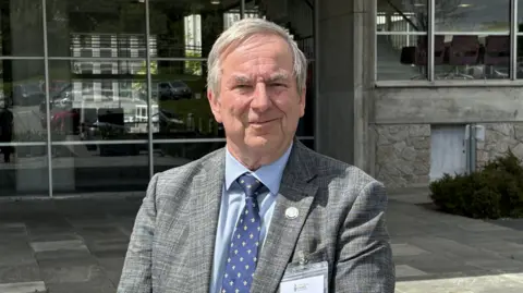 A man with grey hair in a grey jacket with a blue shirt and dark blue tie standing in front of County Hall in Truro