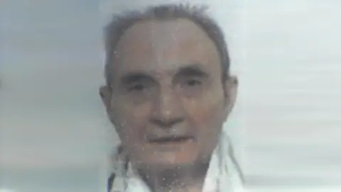 An old analogue photo, possible from a passport or other ID, of a man looking at the camera against a white background. He has short, swept-back greying hair and is wearing a white shirt.