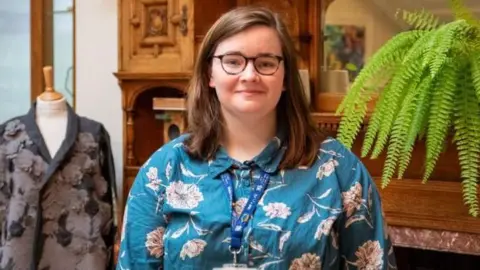 Aine McMenamin stands in a green and beige floral shirt, with a staff lanyard around her neck against a wooden fire surround. There is a mannequin displaying a textile design and a green fern plant behind.