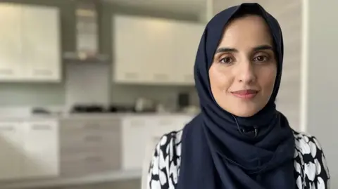 A woman is smiling at the camera and has kitchen units behind her. She is wearing a dark head covering and a black and white top. 