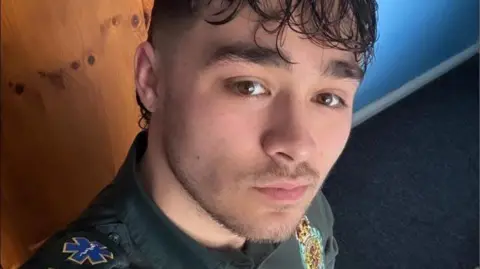 Handout Nico looks into the camera, lips pursed. He has brown eyes and hair and is wearing a green ambulance uniform.