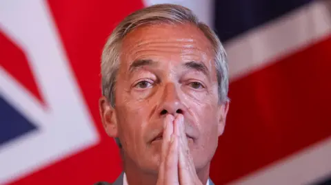 Reuters Nigel Farage with his hands clasped in front of his face. A Union Jack is behind him.