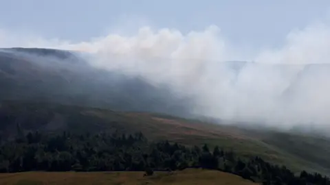 The trees are surrounding the Dovestone Reservoir in Manchester, a large amount of smoke is surrounding the hills in the distance.