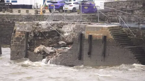 This picture shows a damaged harbour wall. A large section of the stone wall has collapsed, exposing rubble and debris inside. Water from the sea is flowing through the broken area, indicating structural failure likely caused by strong waves, erosion, or storm damage. There are metal safety barriers and fencing around the damaged section. In the background, there are parked cars and buildings. The water in the foreground looks rough.