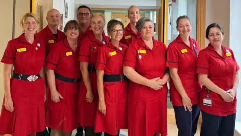 Nine matrons and executive director of nursing lined up in a row in their red uniforms in the hospital corridor.