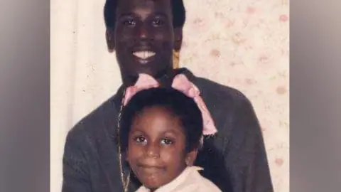 Merseyside Police Jeffrey McNish, who has black hair and wears a grey jumper, smiles at the camera while his sister Kimberley, six, who has pink bows in her haid, sits on his lap 