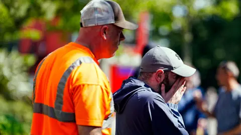 PA Media A man in a grey baseball cap and blue coat looks towards the tributes left for Diogo Jota with his hands pressed agains his face. Another man with an orange high-visibility t-shirt and a brown and white cap stands close behind him with a solemn look on his face