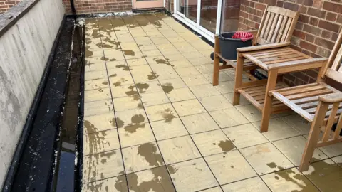 Darren Rozier/BBC A paved balcony area with two wooden seats, following a clean. There is water still drying on the cream-coloured slabs.