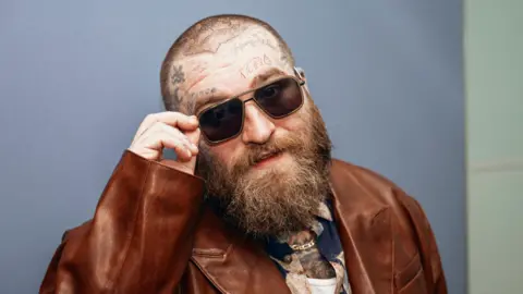 Sarah Louise Bennett/BBC Teddy Swims, wearing a brown leather jacket, points his sunglasses at the camera as he stands in the BBC's new Broadcasting House in 2024.